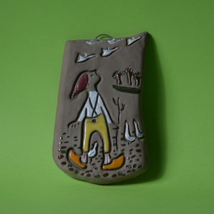 Pode incluir: Um pendurado de parede de cerâmica com fundo castanho e uma cena pintada de uma pessoa com calças amarelas e camisa branca olhando para cima para um bando de pássaros brancos. A pessoa está segurando um pau com um garfo na ponta. Também há pequenas árvores e pássaros pintados no pendurado de parede.