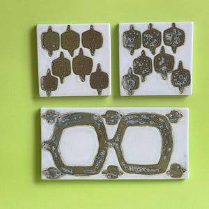 Pode incluir: Três azulejos de cerâmica branca com um padrão geométrico em ouro. Os azulejos têm uma superfície texturizada e estão dispostos em uma fila.