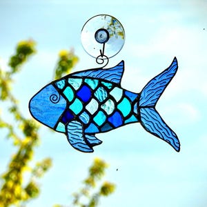 May include: A stained glass fish suncatcher in shades of blue, turquoise, and white. The fish has a detailed scale pattern and is suspended from a suction cup. Decorative art for windows.