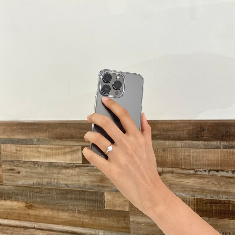 Pu&ograve; includere: Una mano che tiene un iPhone grigio con una custodia trasparente. Il telefono ha un obiettivo della fotocamera con quattro cerchi neri.