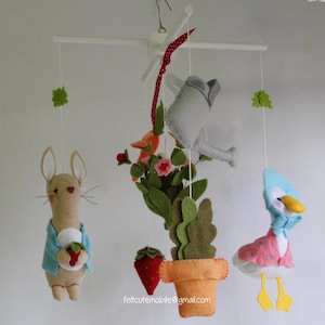 May include: A felt baby mobile with a bunny holding a strawberry, a duck wearing a bonnet, a potted plant with a watering can, and green leaves. The mobile is hanging from a white wooden frame.