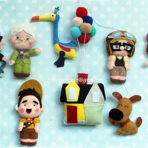 Op de afbeelding: Een vilt mobiel met personages uit de film Up. De mobiel bevat een geel huis, een hond, een vogel, een meisje met rood haar, een jongen in een scoutuniform, een man met een bril, een vrouw met een bril en een bos kleurrijke ballonnen.