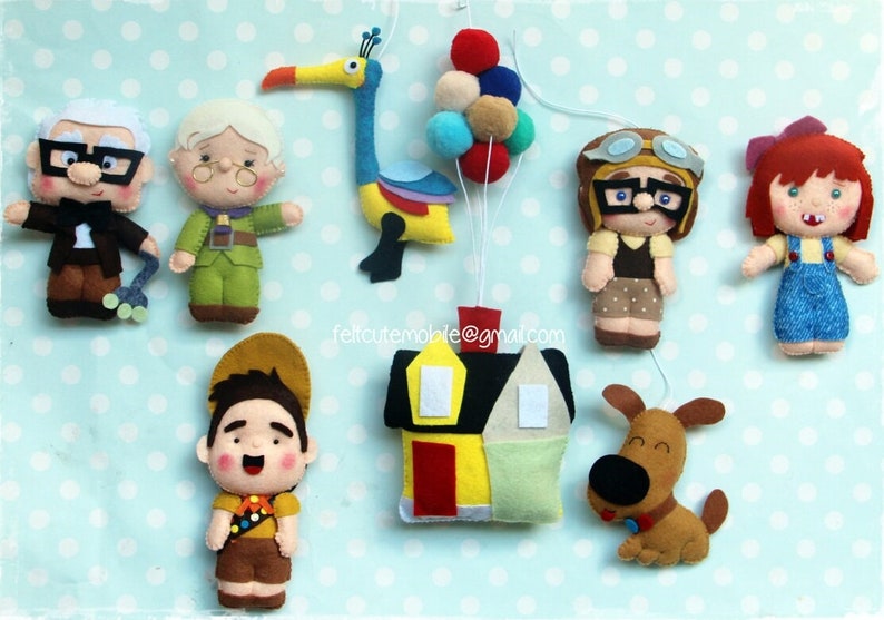 Op de afbeelding: Een vilt mobiel met personages uit de film Up. De mobiel bevat een geel huis, een hond, een vogel, een meisje met rood haar, een jongen in een scoutuniform, een man met een bril, een vrouw met een bril en een bos kleurrijke ballonnen.