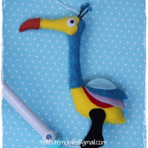 Pode incluir: Um p&aacute;ssaro de feltro amarelo, azul e vermelho com um pesco&ccedil;o comprido e um bico preto. O p&aacute;ssaro est&aacute; pendurado em um cord&atilde;o branco. O fundo &eacute; um padr&atilde;o de bolinhas azuis e brancas.