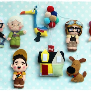 Op de afbeelding: Vilt speelgoedfiguren uit de film Up, waaronder Carl en Ellie Fredricksen, Russell, Dug de hond, Kevin de vogel en een huis. De figuren hangen aan een touwtje met ballonnen.