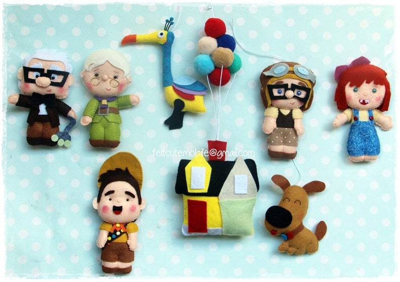 Op de afbeelding: Vilt speelgoedfiguren uit de film Up, waaronder Carl en Ellie Fredricksen, Russell, Dug de hond, Kevin de vogel en een huis. De figuren hangen aan een touwtje met ballonnen.