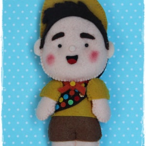 Pode incluir: Um brinquedo de feltro de um menino escoteiro vestindo um chap&eacute;u amarelo, shorts marrons e uma camisa amarela com um remendo colorido. O brinquedo tem um rosto sorridente com bochechas rosadas.