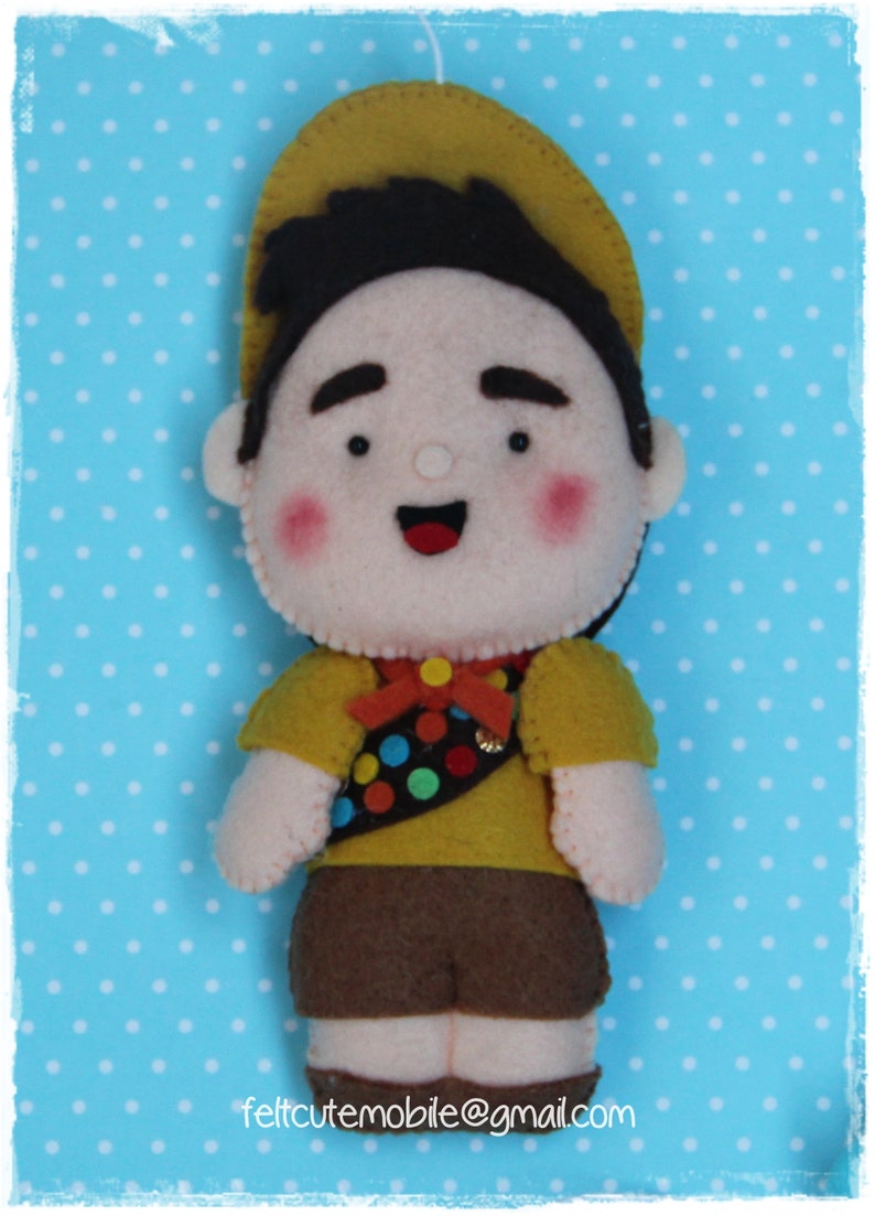 Pode incluir: Um brinquedo de feltro de um menino escoteiro vestindo um chap&eacute;u amarelo, shorts marrons e uma camisa amarela com um remendo colorido. O brinquedo tem um rosto sorridente com bochechas rosadas.