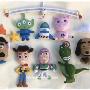 May include: A handmade felt baby mobile featuring characters from the Toy Story movie franchise. The mobile includes Woody, Buzz Lightyear, Rex, Mr. Potato Head, Hamm, Slinky Dog, Alien, and a rocket ship. The mobile is suspended from a white metal frame.