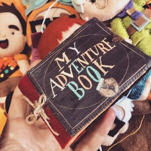Op de afbeelding: Een klein, bruin boek met een rode rand en een gouden lint. Het boek heeft de woorden "MY ADVENTURE BOOK" op de omslag gedrukt in kleurrijke letters.