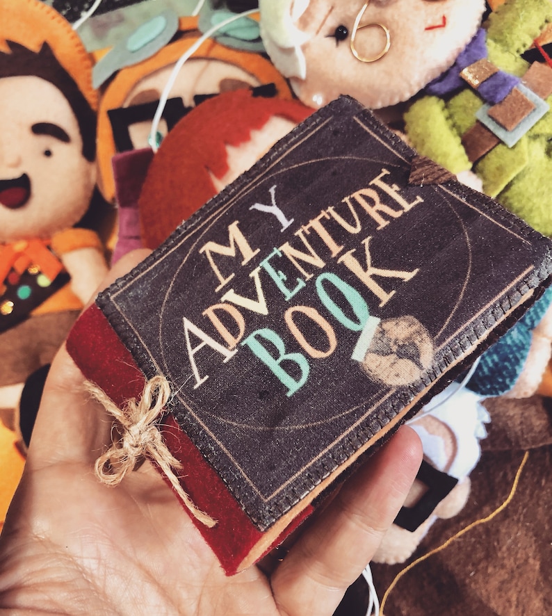 Op de afbeelding: Een klein, bruin boek met een rode rand en een gouden lint. Het boek heeft de woorden "MY ADVENTURE BOOK" op de omslag gedrukt in kleurrijke letters.