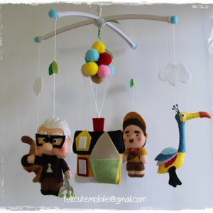 Pode incluir: Um m&oacute;bile de feltro para beb&eacute; com uma casa, uma personagem de Up, um p&aacute;ssaro e um monte de bal&otilde;es. O m&oacute;bile &eacute; branco com uma folha verde de cada lado.