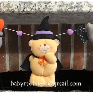 Pode incluir: Uma guirlanda de Halloween com um urso bruxo, um morcego e um caldeirão. A guirlanda é feita de feltro e tem pompons roxos.
