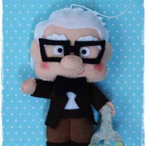 Pode incluir: Uma boneca de feltro de um homem idoso com cabelo branco, usando &oacute;culos, um terno preto e uma gravata borboleta. Ele est&aacute; segurando uma bengala na m&atilde;o direita. A boneca est&aacute; contra um fundo azul com bolinhas brancas.