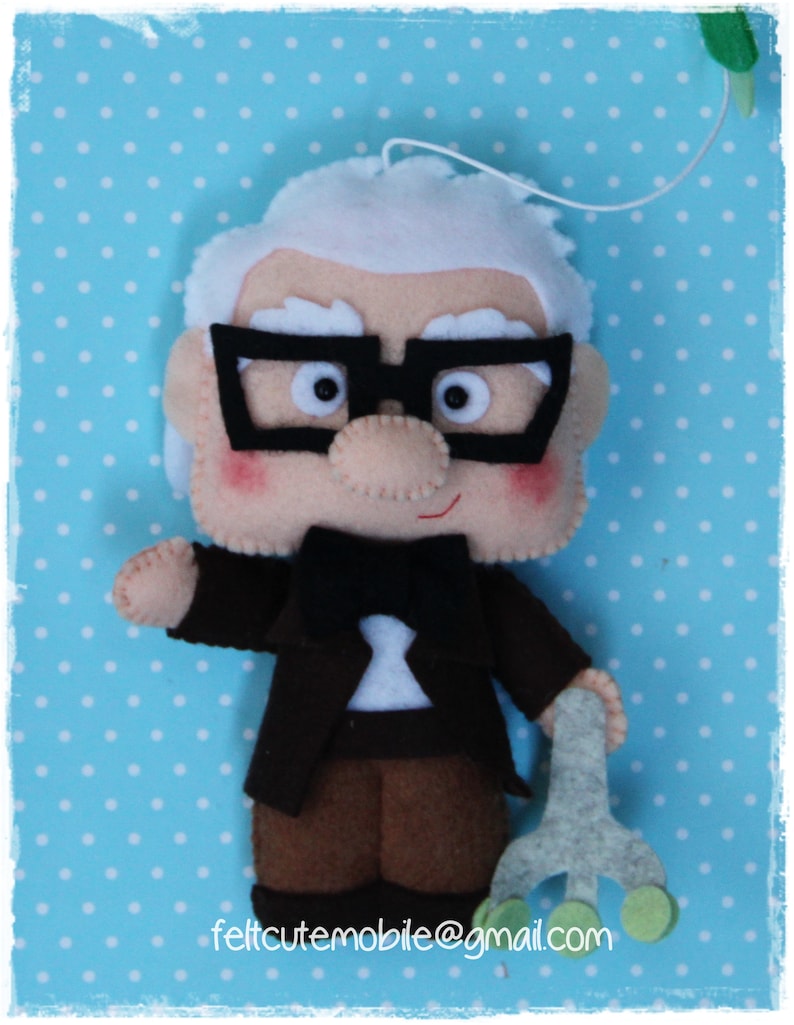 Pode incluir: Uma boneca de feltro de um homem idoso com cabelo branco, usando &oacute;culos, um terno preto e uma gravata borboleta. Ele est&aacute; segurando uma bengala na m&atilde;o direita. A boneca est&aacute; contra um fundo azul com bolinhas brancas.