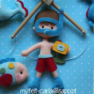 Pode incluir: Um móbile de feltro com um menino usando uma máscara de mergulho azul e nadadeiras, segurando uma câmera amarela. O menino está pendurado em uma haste de madeira. Há também um peixe azul e um peixe amarelo e azul pendurados na haste. O fundo é azul com estrelas brancas.
