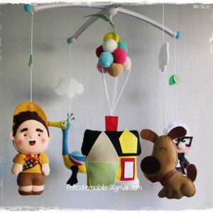 Pode incluir: Um m&oacute;bile de feltro para beb&ecirc; com personagens do filme Up. O m&oacute;bile inclui uma casa, um cachorro, um p&aacute;ssaro e um escoteiro. A casa &eacute; amarela com um telhado vermelho e uma janela branca. O cachorro &eacute; marrom com orelhas pretas e um nariz preto. O p&aacute;ssaro &eacute; azul e amarelo com um pesco&ccedil;o longo e um bico vermelho. O escoteiro est&aacute; vestindo uma camisa amarela, shorts marrons e um chap&eacute;u vermelho. O m&oacute;bile est&aacute; pendurado em um teto branco.