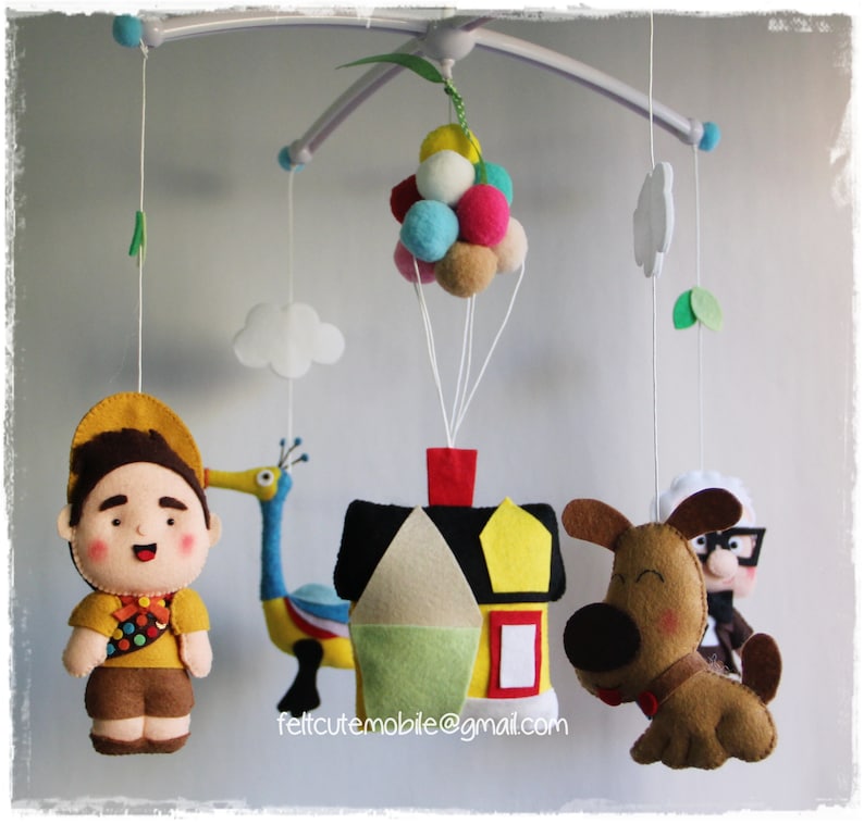 Pode incluir: Um m&oacute;bile de feltro para beb&ecirc; com personagens do filme Up. O m&oacute;bile inclui uma casa, um cachorro, um p&aacute;ssaro e um escoteiro. A casa &eacute; amarela com um telhado vermelho e uma janela branca. O cachorro &eacute; marrom com orelhas pretas e um nariz preto. O p&aacute;ssaro &eacute; azul e amarelo com um pesco&ccedil;o longo e um bico vermelho. O escoteiro est&aacute; vestindo uma camisa amarela, shorts marrons e um chap&eacute;u vermelho. O m&oacute;bile est&aacute; pendurado em um teto branco.