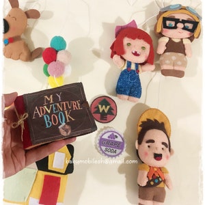 Op de afbeelding: Een vilt mobiel met een boek met de tekst "My Adventure Book", een roodharig meisje met een blauwe overall, een bruine hond, een jongen met een bruine hoed en een scoutuniform, een paarse druivenlimonadeblikje en een groen-wit kompas.