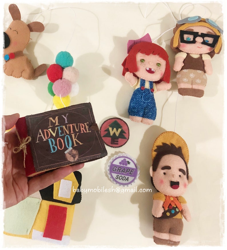 Op de afbeelding: Een vilt mobiel met een boek met de tekst "My Adventure Book", een roodharig meisje met een blauwe overall, een bruine hond, een jongen met een bruine hoed en een scoutuniform, een paarse druivenlimonadeblikje en een groen-wit kompas.
