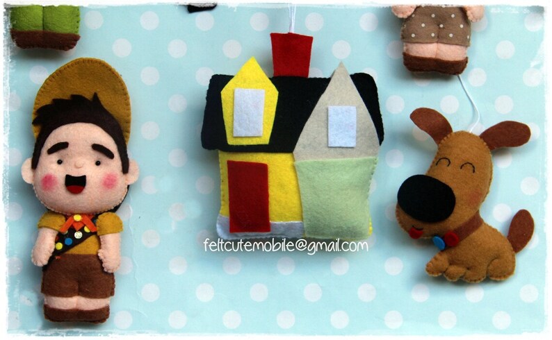 Op de afbeelding: Vilt speelgoedmobiel met een huis, een jongensscout, een hond en de tekst "feltcutemobile@gmail.com".