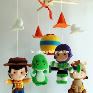Op de afbeelding: Een babymobiel van vilt met een gele en blauwe bal, twee oranje kegels, twee witte wolken, een groene dinosaurus, een paarse en groene ruimtevaarder en een bruine cowboy. De mobiel hangt aan een houten frame.