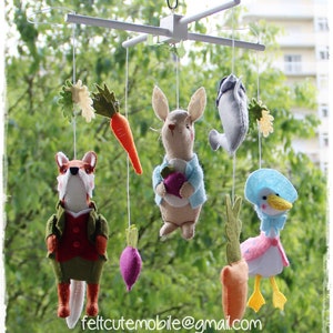 Op de afbeelding: Een witte houten mobiel met vilt dier- en groentedecoraties. De mobiel heeft een vos, een konijn, een eend, een wortel en een raap. De mobiel hangt aan een witte plafondhaak. De tekst "feltcutemobile@gmail.com" is zichtbaar onderaan de afbeelding.