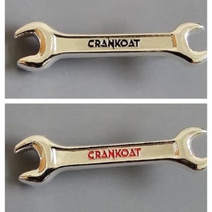 May include: Two silver-colored metal wrench-shaped pins. The top pin features the text CRANKOAT in black lettering, while the bottom pin displays the same text CRANKOAT in red lettering. Both pins have a polished, reflective finish.