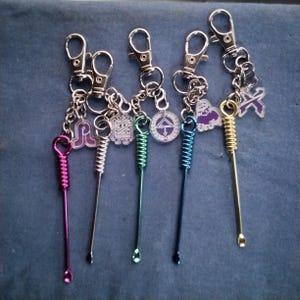 May include: Five metal keychains featuring long, thin stems with coiled tops and small scoops at the bottom. Each keychain includes a silver lobster clasp and a unique acrylic charm in purple and white, showcasing various geometric and graphic symbols.