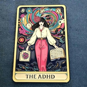 May include: A yellow and black enamel pin with a colorful illustration of a woman with a book and a swirl vortex of colors and objects behind her. The text "THE ADHD" is printed at the bottom of the pin like a tarot card.