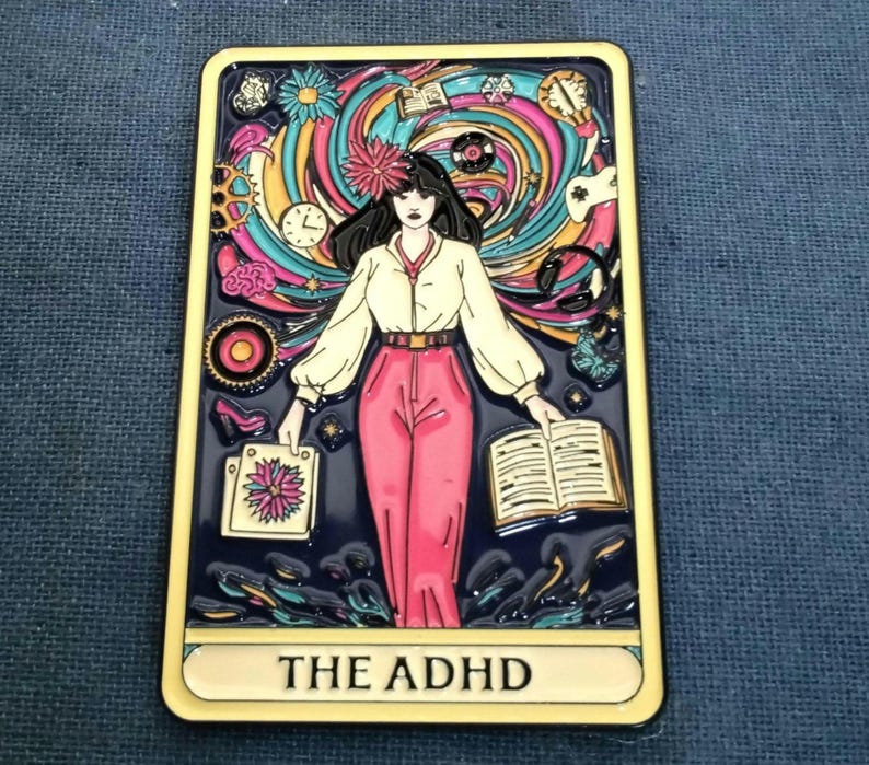 May include: A yellow and black enamel pin with a colorful illustration of a woman with a book and a swirl vortex of colors and objects behind her. The text "THE ADHD" is printed at the bottom of the pin like a tarot card.