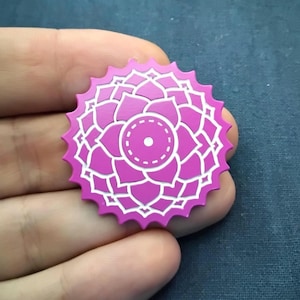 May include: A circular pin featuring a vibrant magenta lotus flower design with white outlines. The emblem has a scalloped, serrated edge and a central circular motif with a small white dot, creating a symmetrical, geometric floral pattern.