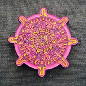 May include: A vibrant, circular pin with a detailed mandala design. The pin features a pink outer ring with yellow accents and a complex pattern of orange, purple, and yellow. The center has a spiral design.