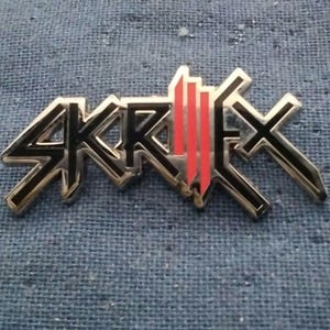 May include: A metal pin featuring the stylized black and red logo of Skrillex. The design consists of sharp, angular lettering with black outlines and a central section composed of three vertical red bars, all set against a metallic silver base.
