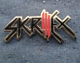 Könnte beinhalten: Eine Metallanstecknadel mit dem stilisierten schwarz-roten Logo von Skrillex. Das Design besteht aus scharfen, kantigen Buchstaben mit schwarzen Umrandungen und einem Mittelteil aus drei vertikalen roten Balken auf einer silbernen Basis.