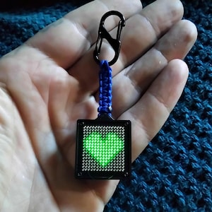 May include: A square keychain featuring a black frame with a glowing green pixelated heart display. The device is attached to a blue paracord lanyard with a braided pattern and a black metal carabiner clip at the top for secure attachment.