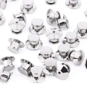May include: A close-up of a group of silver metal pin back closures. The pin backs are round and have a small, raised center. They are used to attach pins, badges, and other items to clothing or fabric.