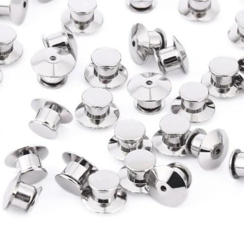 May include: A close-up of a group of silver metal pin back closures. The pin backs are round and have a small, raised center. They are used to attach pins, badges, and other items to clothing or fabric.