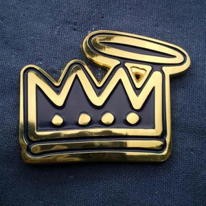 May include: A gold-colored crown pin with a dark blue center and three small white circles. The crown has a halo above it. The pin is set against a dark blue fabric background.