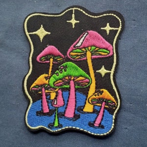 May include: Embroidered patch featuring a cluster of colorful mushrooms with pink, green, and yellow caps against a black background. The patch has a blue border and includes star accents, suitable for jackets, bags, or other fabric items.