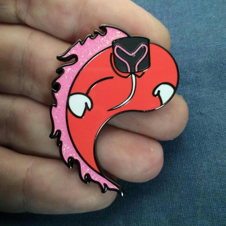 May include: A orange enamel pin with a stylized design. The pin features a orange body with white accents, a black and pink head, and a pink glitter outline. The pin is held in a hand.