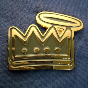 May include: A gold-colored metal pin featuring a crown design with a halo above it. The crown has a detailed, three-dimensional appearance with a dark green background and gold trim. The pin is set against a blue fabric background.