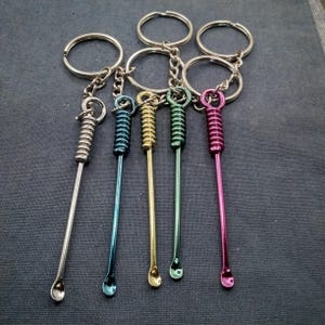 May include: A collection of keychains featuring small, colorful metal tools. Each tool has a loop for the keychain, a coiled section, and a small, rounded scoop at the end. Colors include silver, blue, gold, green, and pink.