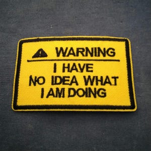 May include: Yellow rectangular patch with black border and text. The patch reads "WARNING I HAVE NO IDEA WHAT I AM DOING" in black capital letters. A black triangle with an exclamation point is above the word "WARNING".