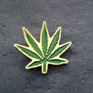 May include: A green and gold enamel pin in the shape of a cannabis leaf. The pin features a detailed design with a gold outline and a green interior. The background is a textured blue fabric.