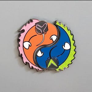 May include: A colorful enamel pin in a yin-yang design. One side is orange with pink glitter accents, the other is blue with green glitter accents. Each side has a white hands and a black and gray helmet.