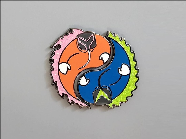 May include: A colorful enamel pin in a yin-yang design. One side is orange with pink glitter accents, the other is blue with green glitter accents. Each side has a white hands and a black and gray helmet.