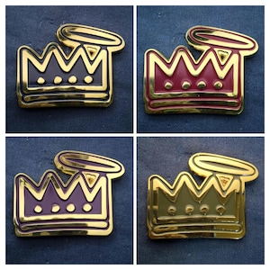 May include: Four gold-colored crown pins with a halo design. Each pin has a different colored crown: black, red, purple, and green. The pins are displayed on a dark blue textured surface.