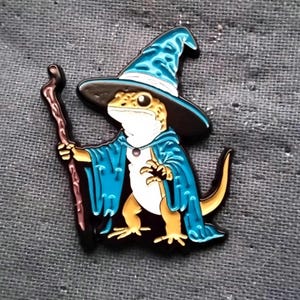 May include: A whimsical enamel pin featuring a lizard dressed as a wizard. The lizard wears a blue robe and a matching pointed hat with a black brim. It holds a brown staff, with a purple gem on the robe.