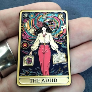 May include: A yellow-colored pin with a colorful illustration of a woman standing in a swirling vortex of colors and objects. The text "THE ADHD" is printed at the bottom of the pin like a tarot card.